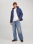 Man draagt een blauwe pufferjas met capuchon, wit T-shirt en wijde jeans.