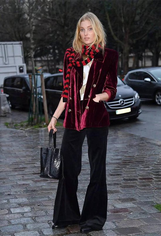 Model in een wijnrode fluwelen blazer met gouden knopen, zwarte wijde broek en een gestreepte sjaal op een stadsplein.