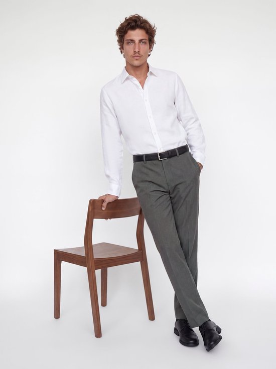 Man met bruin krullend haar en snor, gekleed in een wit overhemd, grijze wijde broek, zwarte riem en zwarte schoenen. Hij leunt tegen een stoel.