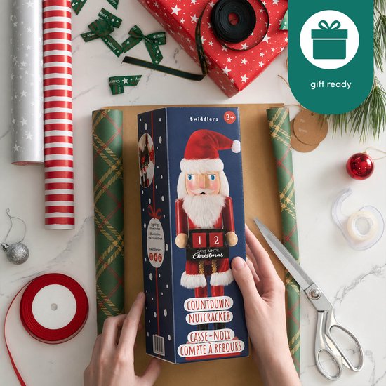 Een kerstman notenkraker verpakt in een blauwe doos, omringd door cadeaupapier en inpakmateriaal. "gift ready" staat erbij.