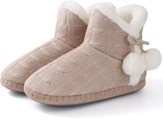 Paar beige gebreide pantoffels met kabelmotief, witte voering van imitatiebont en pompondetails aan de zijkant.