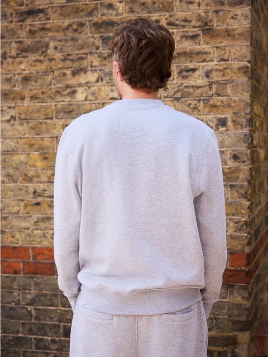 Achteraanzicht van een model in een grijze joggingoutfit, staand voor een bakstenen muur.