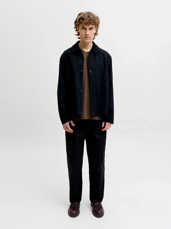 Model draagt een bruin t-shirt onder een zwart jack met kraag en zwarte broek.