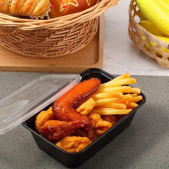 Een zwarte voedselcontainer gevuld met kip, nuggets en frietjes, deksel half open, naast bakkerijproducten in een mand.