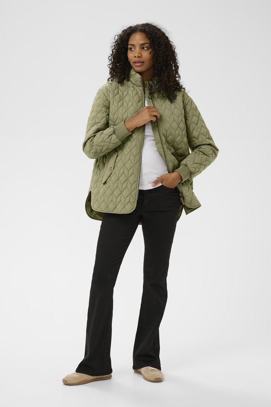 Vrouw met olijfgroene, doorgestikte jas met rits, witte top en zwarte broek.