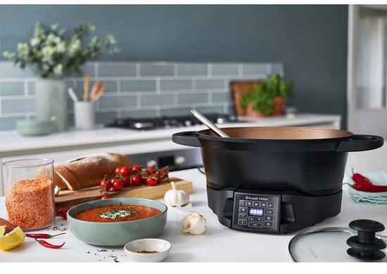 Zwarte multicooker met een digitale display, naast een kom tomatensoep, brood en verse ingrediënten.