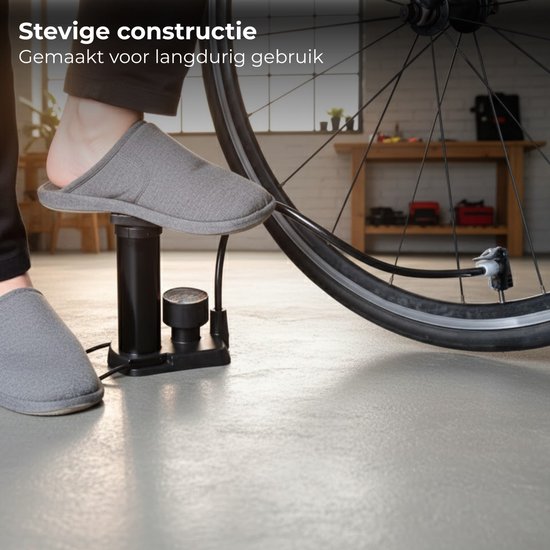 Zwarte fietspomp, bediend met de voet, aangesloten op een fietswiel. Een persoon draagt een grijze pantoffel. Tekst: Stevige constructie. Gemaakt voor langdurig gebruik.