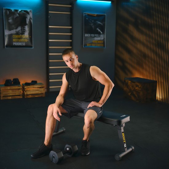 Man met zwarte tanktop en grijze short zit op een zwarte trainingsbank.