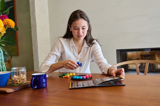 Een jong meisje met een witte blouse speelt het IQ Circle spel aan een houten tafel. Ze houdt een blauw stuk vast en kijkt naar het spelbord.
