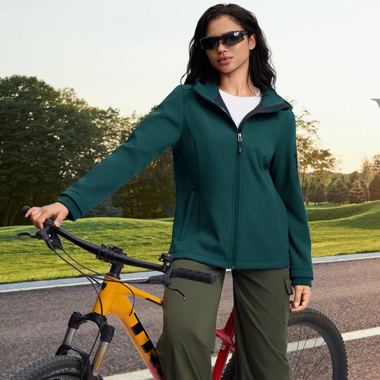 Vrouw met groenblauwe softshell jas en zonnebril staat naast een mountainbike.