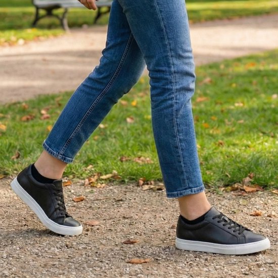 Een persoon draagt zwarte enkelsokken met zwarte sneakers en blauwe jeans, loopt op een grindpad.