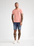 Man met tatoeages in een roze t-shirt en denim shorts met opgerolde pijpen.