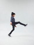 Jongen met zwarte beanie en gestreepte trui in een grijze slim fit spijkerbroek, beweegt met één been omhoog.