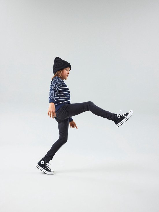 Jongen met zwarte beanie en gestreepte trui in een grijze slim fit spijkerbroek, beweegt met één been omhoog.