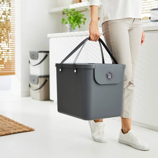 Donkergrijze afvalbak met twee handvatten, gedragen door een persoon in een witte keuken.
