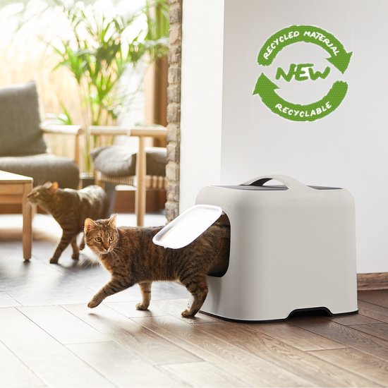 Een rode kat verlaat een lichtgrijze kattenbak in een woonkamer, met een recyclinglogo op de muur.