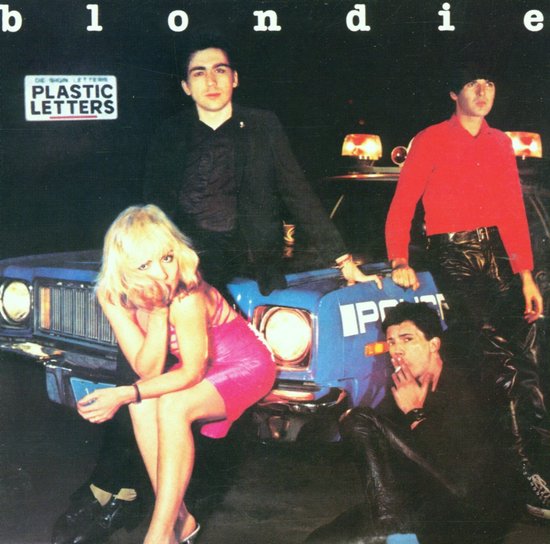 Bandleden van Blondie, inclusief zangeres Debbie Harry in een roze jurk, staan bij een politieauto.