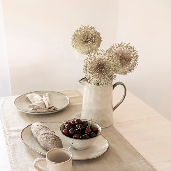 Crèmekleurige kan met bruine rand, gevuld met gedroogde bloemen. Tafel met brood, kersen en servies.