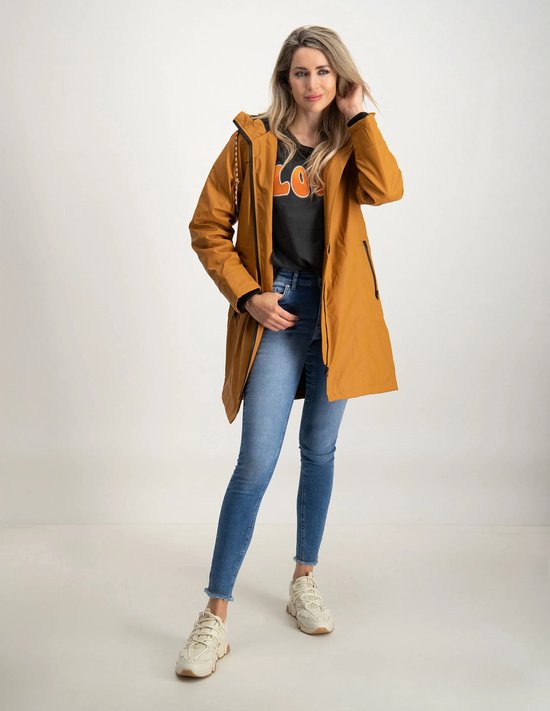 Model in een blauwe skinny jeans met rafelzoom en een okergele jas, gedragen met sneakers.