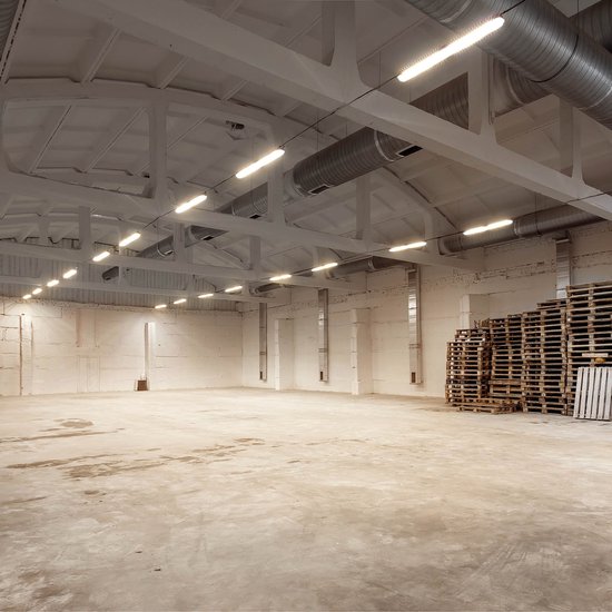 Grote, lege opslagruimte met hoge plafonds, betonnen vloer en tl-verlichting.