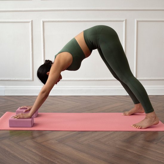 Vrouw in groene sportkleding in neerwaartse hondhouding op roze mat, handen op roze yogablokken.