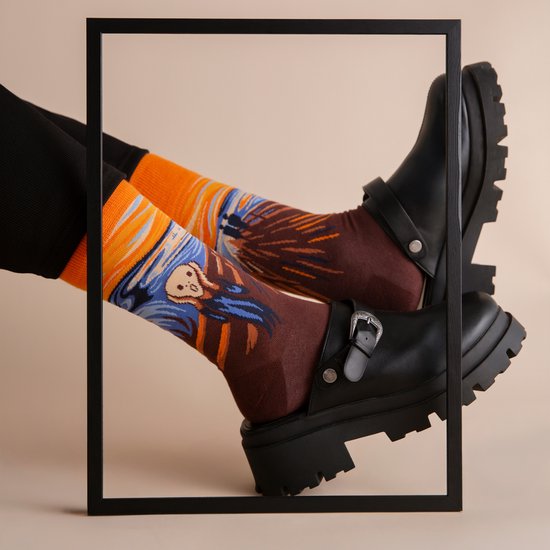 Model draagt zwarte broek, zwarte klomp-sneakers en sokken met Munchs Schreeuw in oranje en bruin.