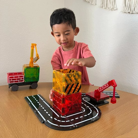 Jongetje speelt met Magna-Tiles. Een hijskraan met een groene lading staat naast een stapel rode en gele blokken op een speelgoedweg.