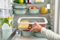 Une main sortant du réfrigérateur une boîte de conservation transparente remplie d'aliments. La boîte est de couleur verte.