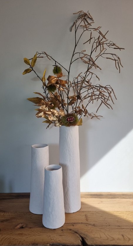 Drie witte, conische vazen met ruw, steenachtig oppervlak, waarvan één gevuld is met bruine takken en herfstbladeren.