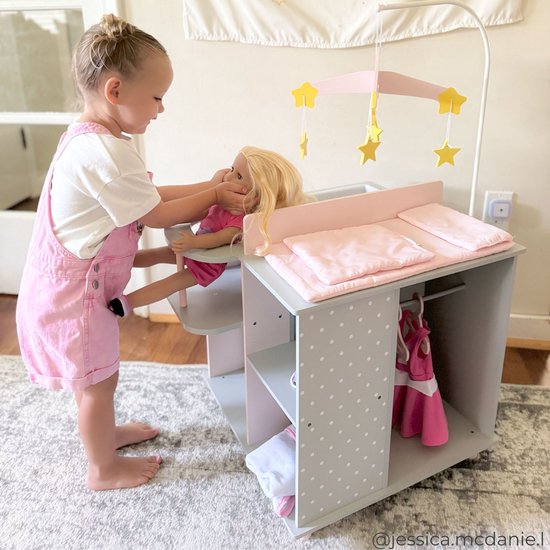Meisje speelt met een pop bij een grijze commode met roze details, witte stippen en een mobiel met gele sterren.