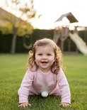 Een blij baby meisje in een roze gebreide trui kruipt op een grasveld buiten, met een speeltoestel op de achtergrond.