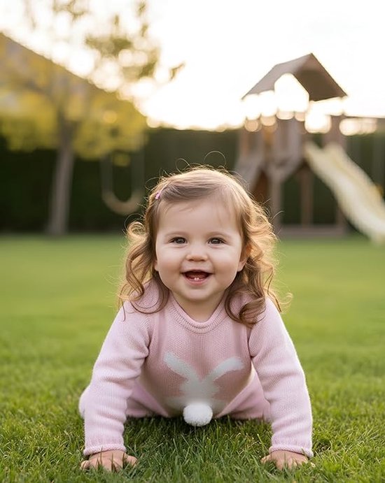 Een blij baby meisje in een roze gebreide trui kruipt op een grasveld buiten, met een speeltoestel op de achtergrond.