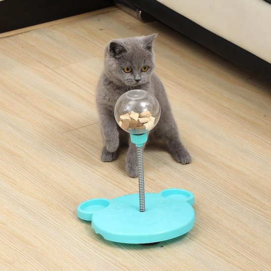 Grijze kat kijkt naar een blauwe verende voerbal met brokjes erin.