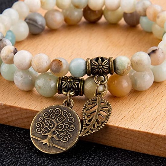 Twee armbanden van lichtgroene, beige en bruine kralen met bronzen bedels van een levensboom en een veertje, op een houten ondergrond.