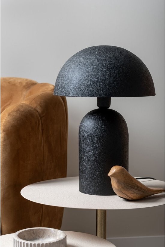 Zwarte tafellamp met ronde voet en bolvormige kap, geplaatst op een bijzettafel naast een okergele fauteuil. Een houten vogel decoratie staat ernaast.