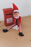 Elf met kerstmuts, rood shirt en gestreepte broek, zittend bij een rode postbus, met een rapport.