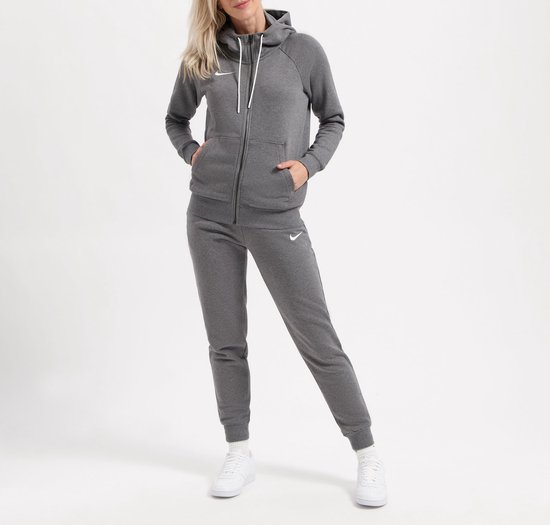 Grijze Nike hoodie met rits, capuchon en witte trekkoordjes, met Nike logo op de borst. De vrouw draagt een joggingbroek en witte sneakers.
