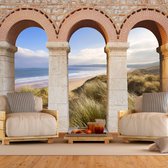 Fotobehangkoning - Behang - Vliesbehang - Fotobehang Uitzicht op het Strand door de Romeinse Pilaren 3D - 300 x 210 cm