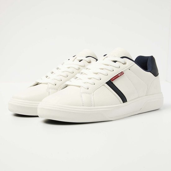 Paar witte sneakers met veters, donkerblauwe accenten op de hiel en zijkant. Rode tag met logo.