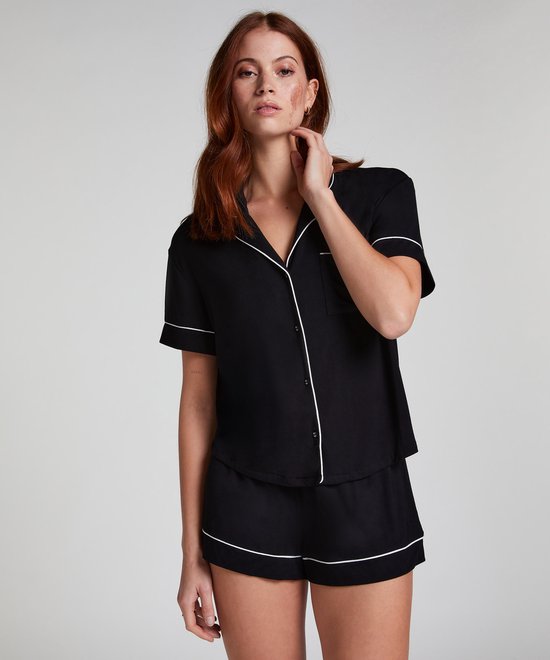 Model met rood haar draagt een zwart pyjamashirt met korte mouwen en witte piping. Het shirt heeft een open kraag en borstzakje.