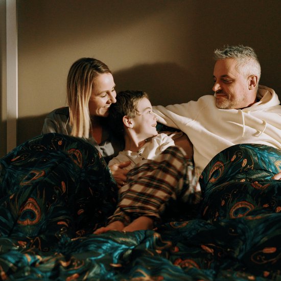 Een gezin, bestaande uit een man, vrouw en zoon, knuffelen samen onder een dekbed met pauwenverenprint.