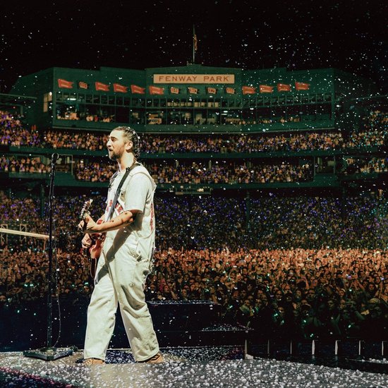 Noah Kahan speelt gitaar op een podium in Fenway Park, met een groot publiek en confetti.