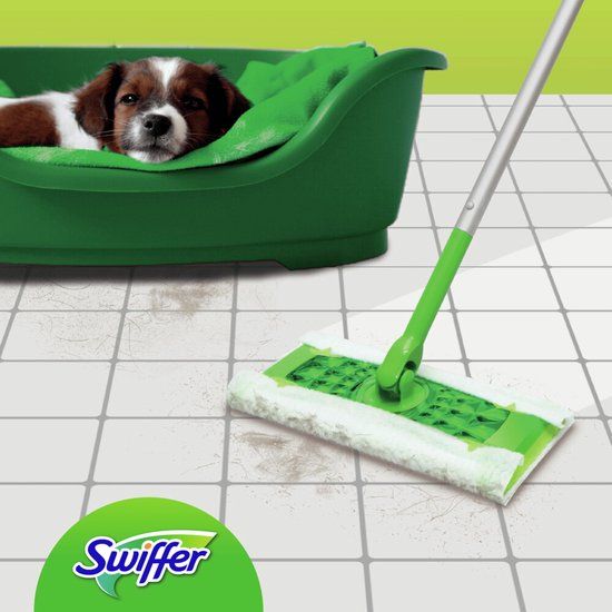 Une serpillière Swiffer avec manche sur un sol carrelé avec des poils de chien. Un chiot est allongé dans un panier vert à l'arrière-plan.