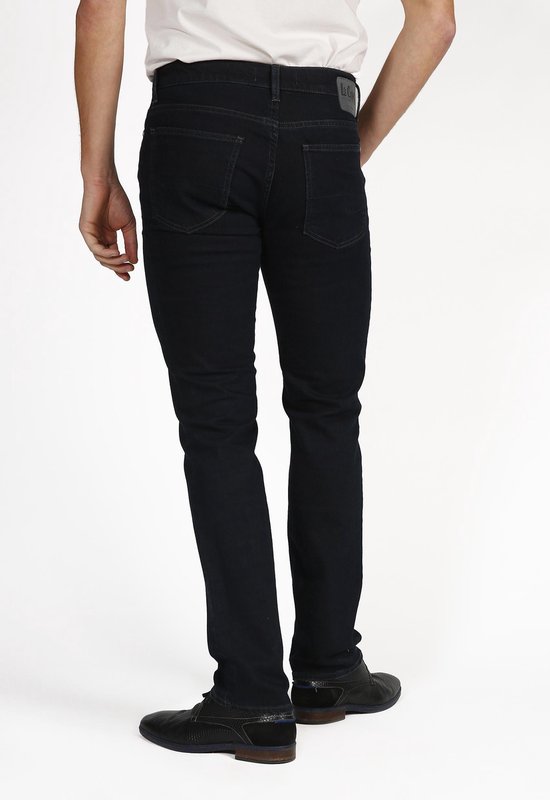 Man in donkere Lee Cooper jeans, staand van achter, witte shirt, donkere schoenen.