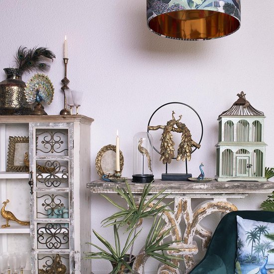 Ornements de paon dorés sur une console, avec des plumes de paon dans un vase, des bougies et une cage à oiseaux vert pâle.