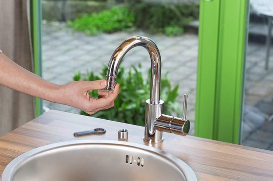 Een hand houdt een grijze metalen schroefdraad adapter bij een glanzende chromen kraan boven een rvs spoelbak.