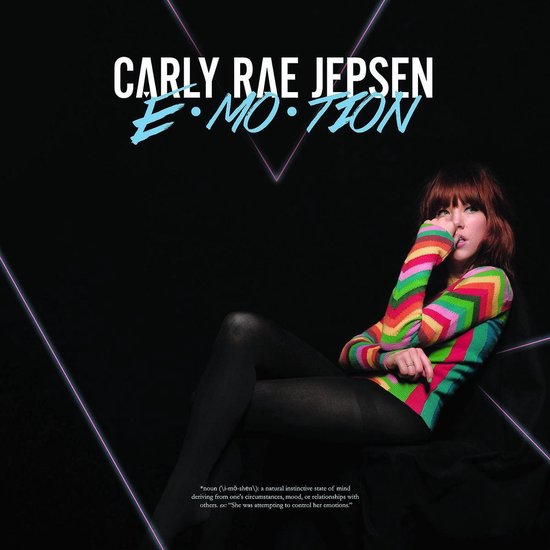 Carly Rae Jepsen in een kleurrijke, zigzag-gebreide trui met regenboogkleuren, zwarte panty's en zittend op een zwarte achtergrond.