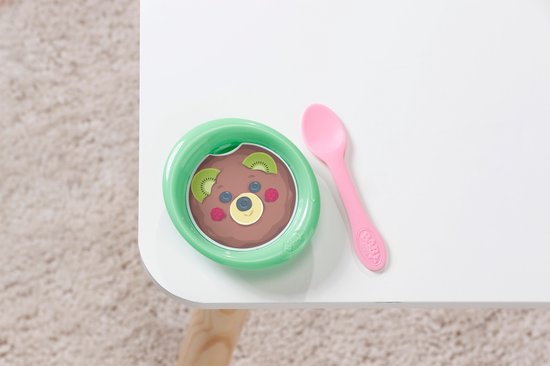 Groen schaaltje met bruine pudding en kiwi, naast een roze lepel op een witte tafel.