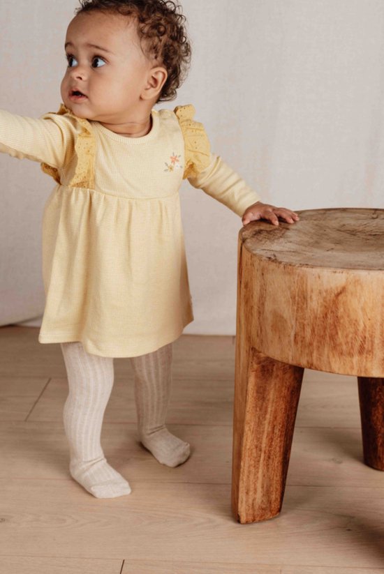 Baby in een gele geruite jurk met lange mouwen, kanten details op de schouders en een bloemenapplicatie.
