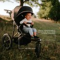 Een lachende baby zit in een zwarte driewielkinderwagen in een natuurlijke omgeving met gras.